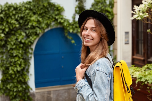 Foto de lado de uma mulher adorável e alegre passeando pela velha rua com vegetação verde, usando um chapéu da moda e jaqueta jeans