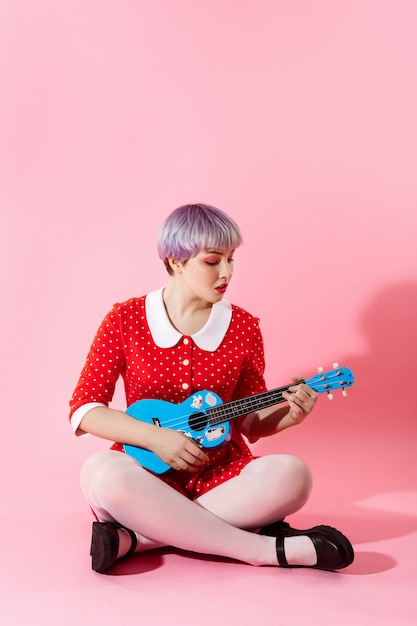 Foto de linda garota dollish com cabelo violeta curto curto, vestido vermelho, tocando ukulele azul sobre parede rosa