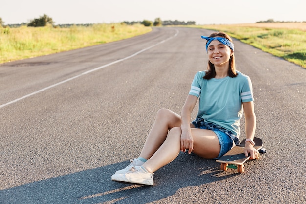 Foto de linda mulher sorridente vestindo camiseta, faixa de cabelo e curta, sentada na rua com longboard, olhando para a câmera com um sorriso, expressando felicidade, aproveitando o tempo de lazer.