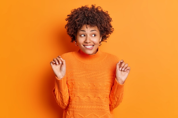 Foto de meio comprimento de mulher afro-americana positiva morde os lábios, mantém as mãos levantadas, olha curiosamente para longe, sorrisos intrigados, percebe que algo interessante usa um macacão casual isolado sobre uma parede laranja