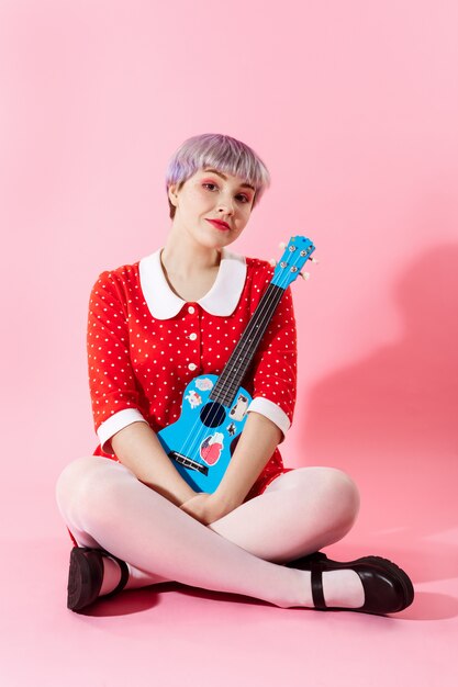 Foto de menina bonitinha bonita com cabelo violeta claro curto, vestido vermelho, segurando o ukulele azul sobre parede rosa
