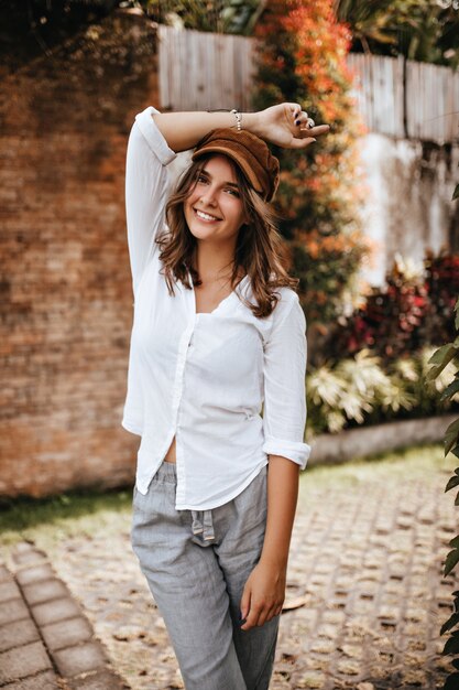 Foto de menina com cabelo curto, usando boné marrom, blusa branca e calça de linho, que ergueu a mão contra o espaço de um prédio de tijolos e arbustos.