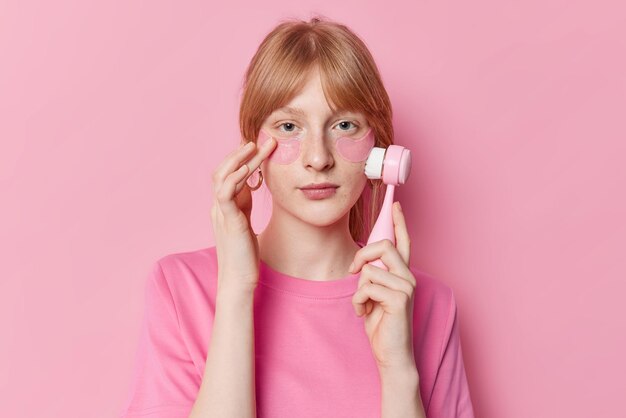 Foto de menina de quinze anos de cabelos ruivos atenta cuida da pele facial usa escova de massagem aplica almofadas de hidrogel sob os olhos usa camiseta casual isolada sobre fundo rosa Conceito de bem-estar