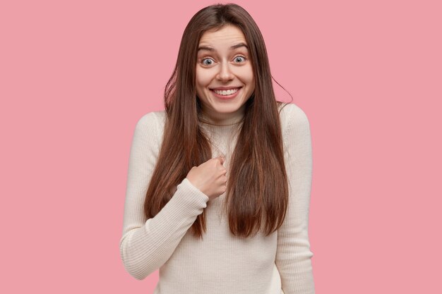 Foto de moça morena alegre aponta para si mesma, tem sorriso dentuço, pergunta sobre alguma coisa, sendo escolhida