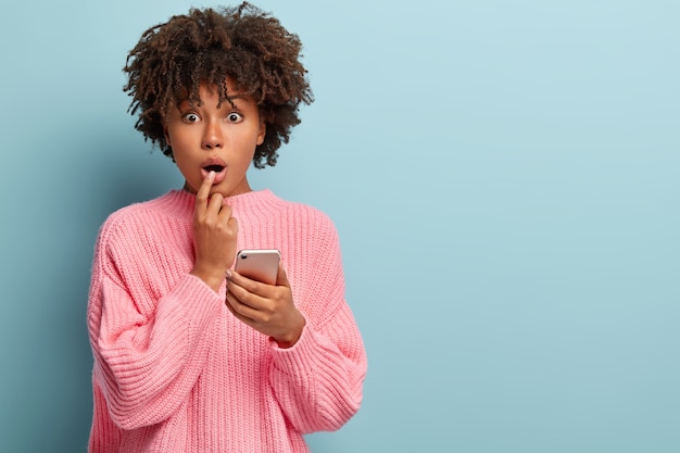 Foto de mulher afro-americana atônita fazendo compras online, segura o celular, fica de queixo caído, expressa surpresa, vestida com um macacão de malha solto, recebe uma notificação estranha de um amigo.