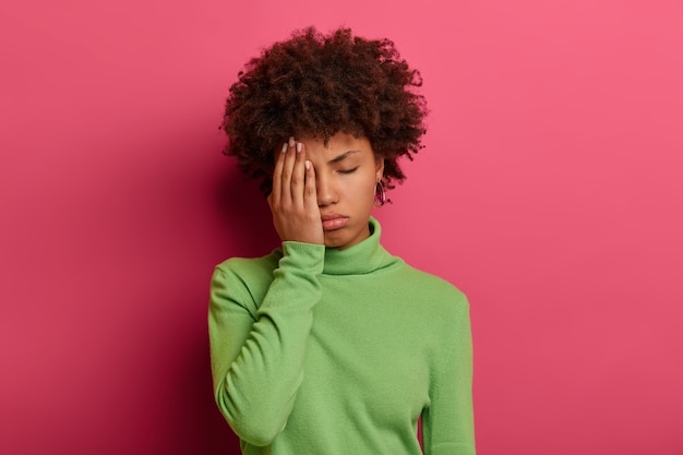 Foto de mulher afro-americana cansada cobre metade do rosto com a palma da mão, mantém os olhos fechados, quer dormir, precisa descansar