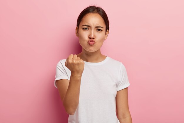 Foto de mulher asiática irritada e insatisfeita fecha o punho em desgosto, mantém os lábios fechados, faz uma expressão de indignação no rosto, usa roupas brancas, ameaça você, isolada na parede rosa.