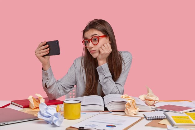 Foto de mulher atraente segura um celular moderno na frente do rosto, faz videochamada, trabalha como freelance em casa e prepara relatório criativo