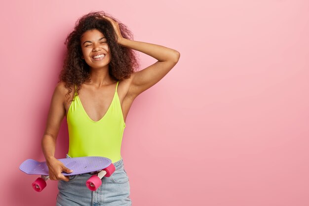 Foto de mulher bonita com cabelos crespos, tocando a mão, tem sorriso agradável, veste roupa de verão, segura skate roxo e rosa