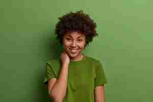 Foto grátis foto de mulher bonita com pele perfeita e sorriso cheio de dentes, toca o pescoço, estar de bom humor, tem conversa casual, usa camiseta verde verão, poses internas. conceito de emoções humanas positivas