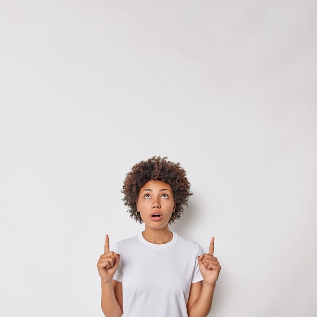 Foto de mulher bonita de cabelos encaracolados aponta acima com os dois dedos indicadores em cima mostra propaganda incrível usa camiseta básica casual isolada sobre fundo branco do estúdio omg, olhe lá