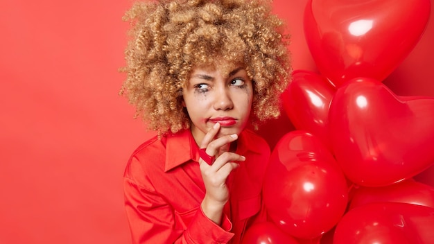 Foto de mulher encaracolada descontente vazou maquiagem depois de chorar segura balões de coração parece mal-humorado olha para longe usa poses de camisa contra fundo vermelho espaço em branco no lado esquerdo Conceito de celebração