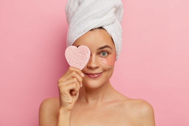Foto de mulher jovem aplica adesivos sob os olhos para ter uma pele fresca e aparência jovem, cobre os olhos com uma esponja cosmética, usa uma toalha branca na cabeça após tomar banho, cuida da beleza