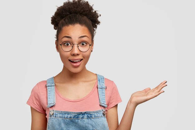 Foto de mulher jovem e atraente surpreendeu a expressão satisfeita, levanta a palma da mão contra a parede branca, usa óculos e roupa casual, tem pele escura.