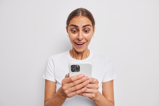 Foto de mulher morena lendo mensagem recebida com respiração suspensa olhando para a tela do celular é incrível oferta fica emocionada tem olhos saltados usa camiseta casual isolada na parede branca