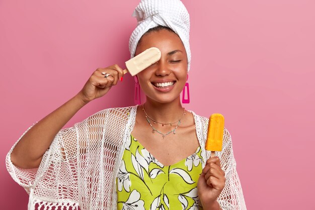 Foto de mulher morena positiva se diverte e segura delicioso sorvete frio, cobre os olhos com picolé, tem sorriso largo, vestida com roupa doméstica, isolada na parede rosa. Verão, alegria, comer