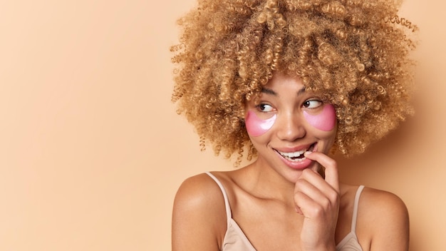 Foto de mulher sonhadora satisfeita morde o dedo desvia o olhar com expressão pensativa aplica remendos de hidrogel rosa para tratamento de pele isolado sobre espaço de maquete de fundo bege para sua promoção