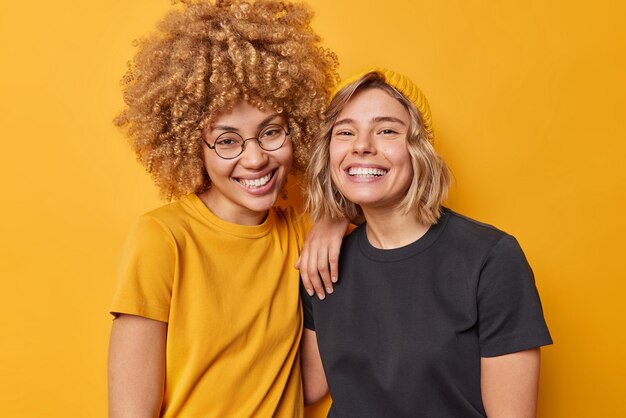 Foto de mulheres bonitas e alegres ficam ao lado uma da outra vestidas com camisetas casuais sorriem alegremente isoladas sobre o fundo amarelo vívido do estúdio Pessoas amizade conceito de emoções positivas