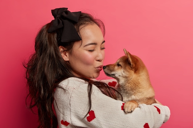 Foto de perfil de mulher adorável abraça e beija cachorro com amor, não via animal de estimação há muito tempo, gosta de brincar com animais, isolado sobre o espaço rosa. momento romântico