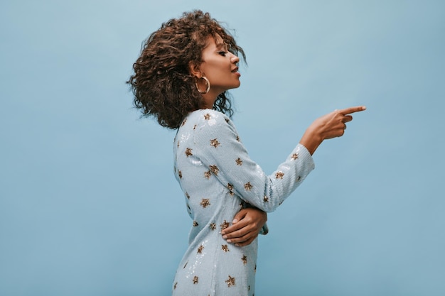 Foto de perfil de mulher na moda com cabelos ondulados em vestido azul moderno e brincos mostrando para colocar texto em fundo isolado