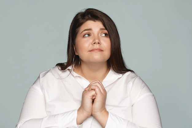 Foto de preocupada frustrada e esperançosa jovem e gorda, com cabelo escuro e corpo curvilíneo, mantendo as mãos juntas, olhando para cima, orando a deus, seus olhos cheios de fé e esperança