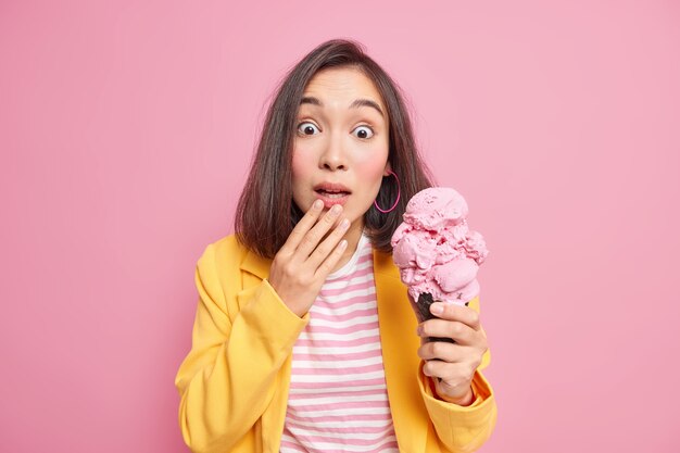 Foto de surpresa jovem asiática com cabelo escuro olha chocada vestida com camiseta listrada e jaqueta amarela contém saboroso sorvete isolado sobre a parede rosa. Conceito de pessoas e emoções