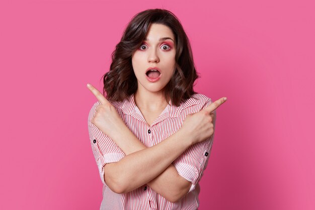 Foto de surpresa jovem europeu com maquiagem, cruza as mãos no peito, abre a boca, vestida com camisa listrada, isolada em rosa. Pessoas, propaganda, conceito de reação.