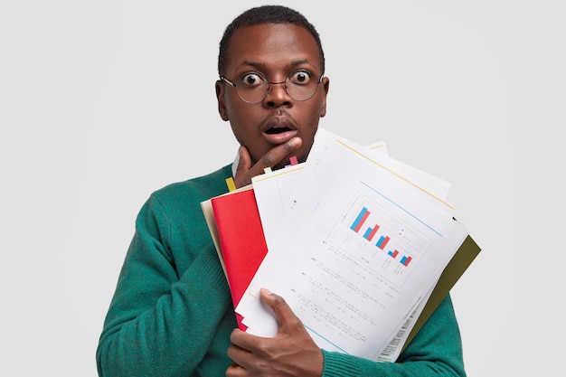 Foto de um cara de pele escura estupefato com as mãos no peito, analisa informações de documentos em papel, estuda dados de informações