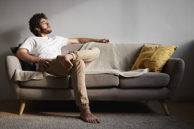 Foto de um cara jovem e elegante com barba crespa, penteado volumoso e pés descalços, de olhos fechados, adormecendo ou ouvindo música clássica, aproveitando os momentos de lazer, sentado no sofá