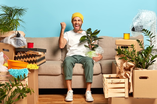 Foto de um cara muito feliz com os punhos cerrados, posando no sofá, segurando a planta de casa embrulhada