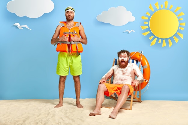 Foto de um cara ruivo surpreso com protetor solar na pele queimada de sol, repousa na cadeira de praia, olha com choque para a câmera, amigo está perto, usa colete salva-vidas protetor, óculos de proteção. Dois companheiros na praia