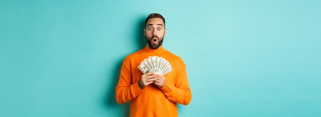 Foto grátis foto de um cara surpreso segurando dinheiro parecendo espantado com dólares contra fundo turquesa