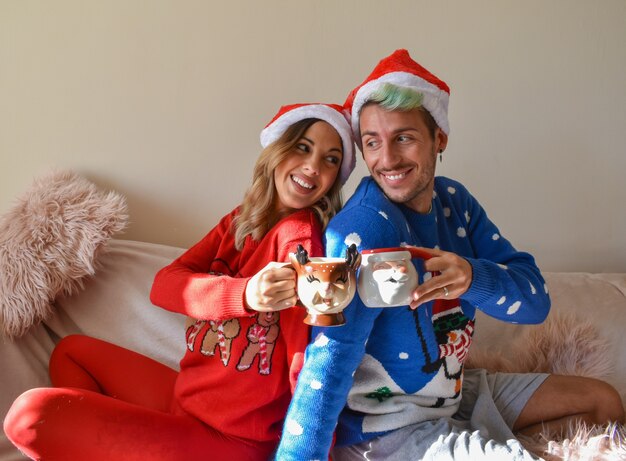 Foto de um casal feliz em cabanas de natal e roupas segurando xícaras engraçadas