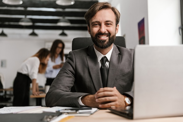 Foto de um empresário alegre em um terno formal, trabalhando com documentos e laptop, enquanto está sentado à mesa em um escritório de plano aberto