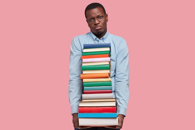 Foto de um empresário de pele escura descontente com uma pilha de livros científicos, uma expressão facial infeliz, uma camisa formal e óculos, isolados sobre a parede rosa do estúdio