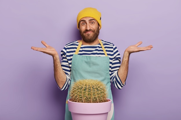 Foto de um florista masculino hesitante espalha as palmas das mãos para os lados, tem uma expressão facial desavisada, usa um uniforme especial, isolado sobre um fundo roxo. Conceito de botânica