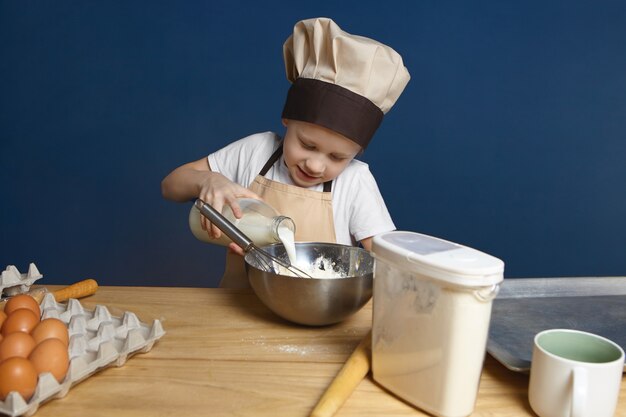 Foto de um garotinho alegre de avental e boné cozinhando sobremesa em um grande balcão de madeira com ovos