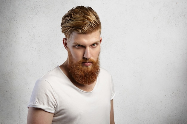 Foto de um hippie barbudo autoconfiante com corte de cabelo estiloso e constituição musculosa, vestindo uma camiseta com mangas enroladas, parecendo com expressão severa, zangado com algo.