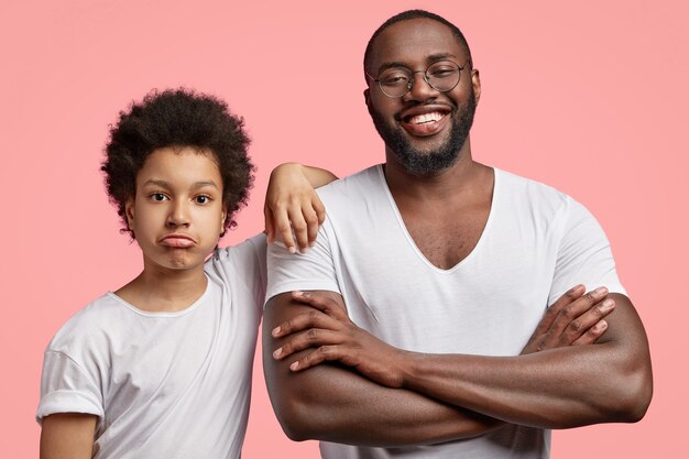 Foto de um homem adulto negro e sorridente satisfeito com um sorriso brilhante, mantém os braços cruzados, passa o tempo livre com o filho