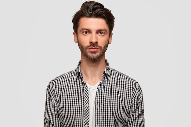 Foto de um homem atraente com um penteado elegante, tem barba por fazer, parece diretamente sério, usa uma camisa quadriculada, isolada sobre uma parede branca. Gerente confiante do sexo masculino trabalha, modelos internos