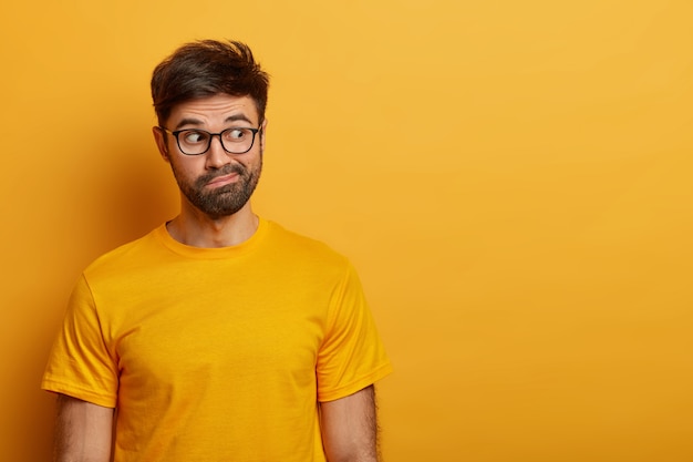 Foto de um homem barbudo hesitante olha de lado, sorri afetado e tem uma expressão intrigada, tenta decidir algo, vestido com uma camiseta amarela casual, posa sobre uma parede vibrante, se perguntando o que vê