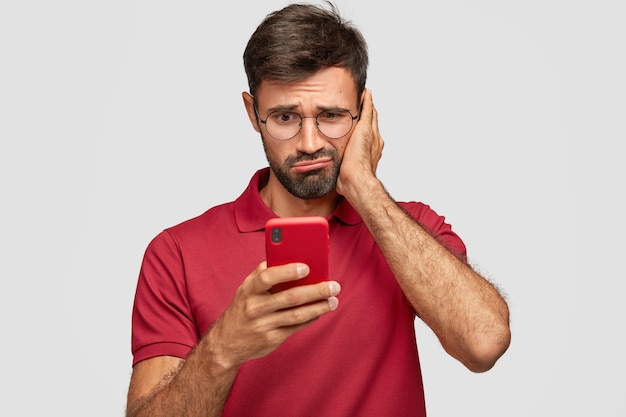 Foto grátis foto de um homem barbudo insatisfeito segura um celular moderno, lê notícias negativas on-line no site, recebe mensagem de lembrete de pagar contas, usa roupas vermelhas, isolado sobre uma parede branca