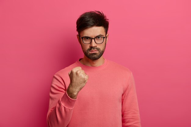 Foto de um homem bravo com a barba cerrada o punho, parece com irritação, promete punir o colega por estar atrasado, usa roupas casuais, posa contra a parede rosa brilhante.