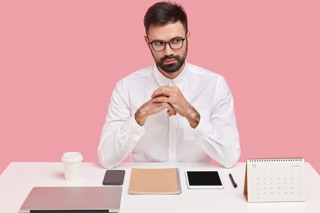 Foto de um homem de negócios bonito e barbudo tem uma expressão atenciosa, mantém as mãos juntas, vestido com roupa formal, tem tudo em seus lugares na mesa