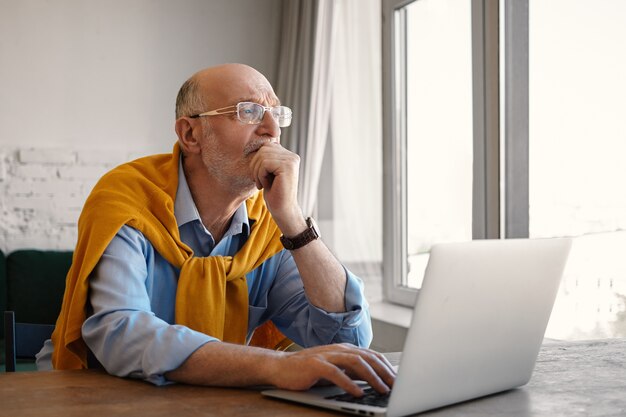 Foto de um homem de negócios sênior atraente e elegante de setenta anos de idade usando óculos e roupas formais, com uma aparência pensativa e pensativa, enquanto trabalhava em um laptop, sentado à janela