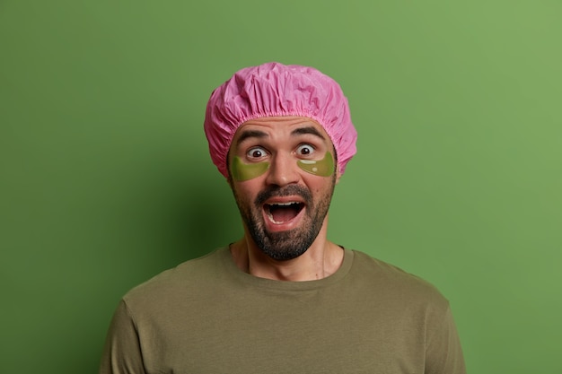 Foto de um homem emocional assustado com os olhos arregalados e gritos de medo, aplica almofadas de colágeno sob os olhos, faz tratamentos de beleza, usa chapéu de banho protetor, fica em ambientes fechados. bem estar beleza