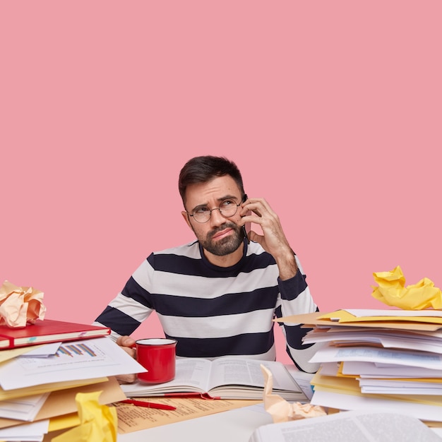 Foto de um homem europeu com a barba por fazer que confunde a expressão facial, fala pelo telefone celular, usa um macacão casual preto e branco, bebe café