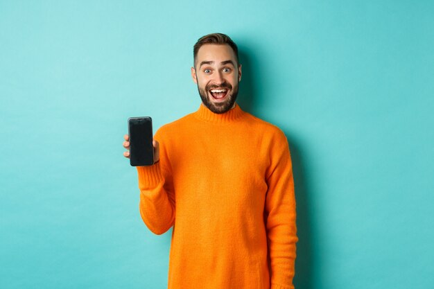 Foto de um homem feliz, mostrando a tela do celular, apresentar a loja online, aplicativo, em pé sobre a parede turquesa.