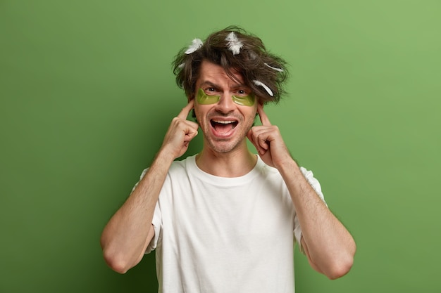 Foto grátis foto de um homem irritado tapa os ouvidos e grita, exige silêncio, acordei com muito barulho, usa camiseta casual branca, manchas hidratantes sob os olhos, tem mau humor pela manhã, isolado na parede verde