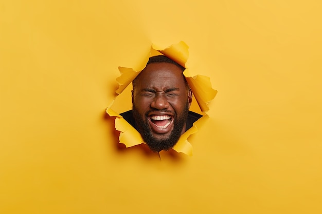 Foto de um homem negro com a barba por fazer ri de alegria, se sente entretido, posa em um fundo amarelo rasgado, tem um sorriso cheio de dentes, dentes brancos